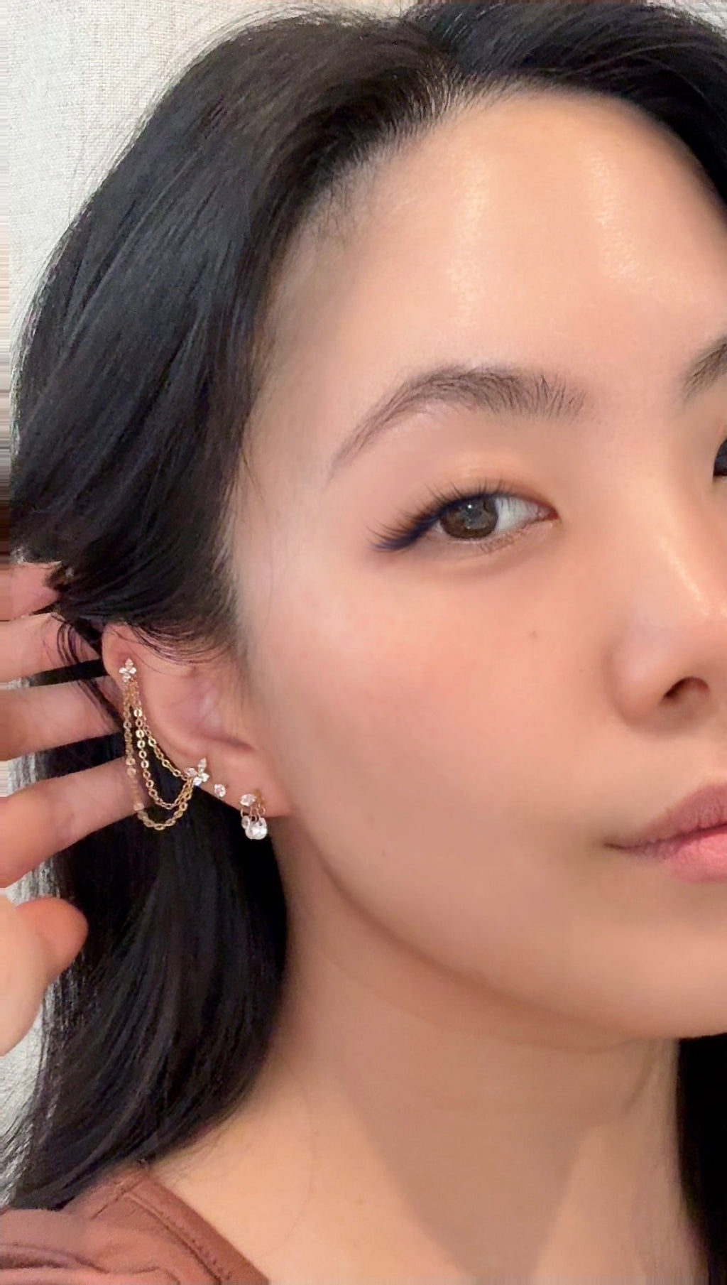A close-up of a woman wearing a delicate gold ear cuff with small dangling charms and clear rhinestone accents, with her hand adjusting the earring.
