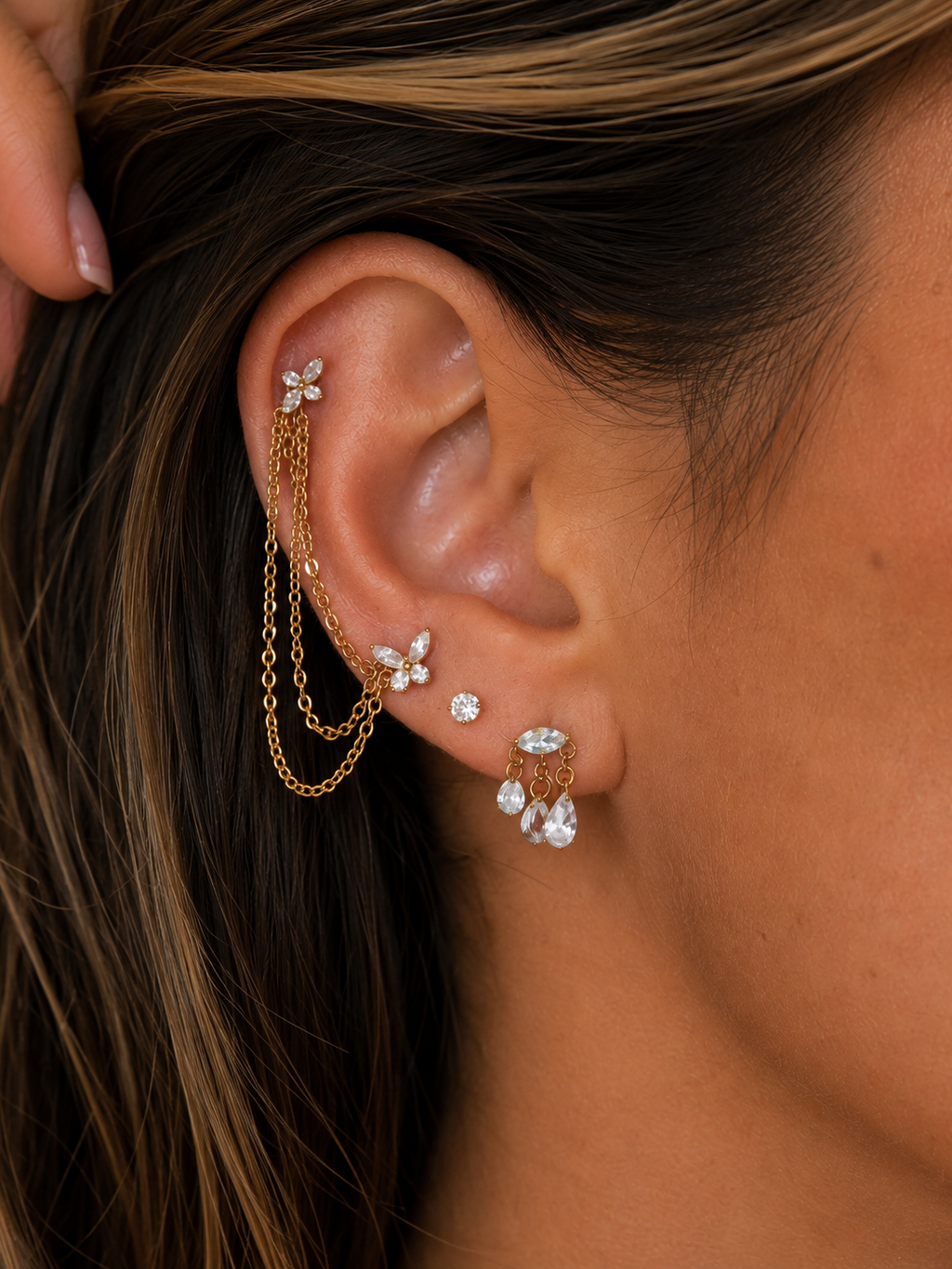 A close-up of a model's ear showcasing a multi-piece gold ear cuff set adorned with clear crystals. The set includes a flat ear cuff with hanging chains, butterfly studs, a small stud, and a cluster drop earring.